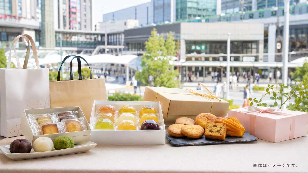 浦和駅周辺の明るい駅前風景を背景に、上品な和菓子、フルーツゼリー、焼き菓子のギフト箱、買い物袋を自然に配置した実写風写真