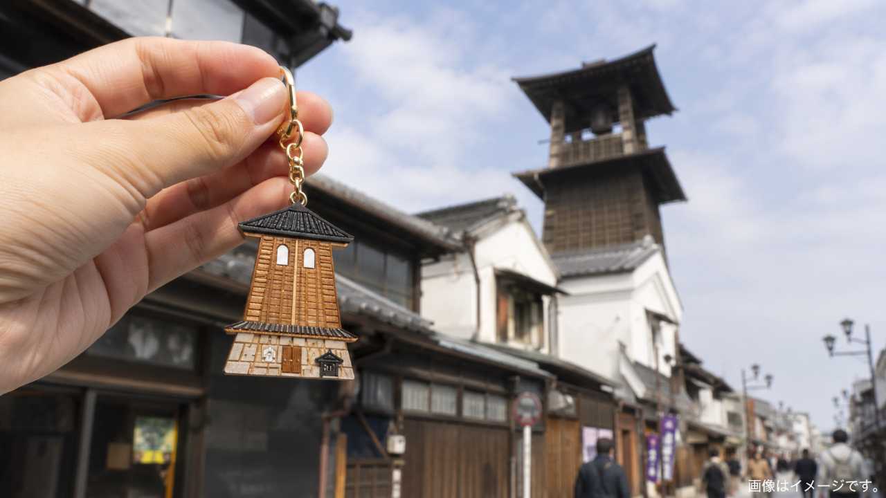 川越の蔵造りの町並みと時の鐘を背景に、小江戸風のかわいいキーホルダーを手に持っている写真風アイキャッチ