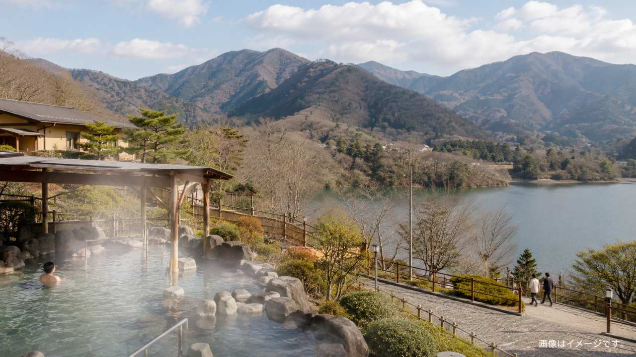 飯能市の山並みと湖畔を背景に、湯けむりが立つ温泉施設を遠景で描いた実写風