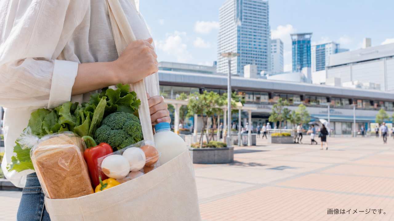 埼玉県の大宮駅周辺をイメージした都市の駅前風景と、エコバッグに野菜、牛乳、パン、卵を入れて買い物を終えた人物