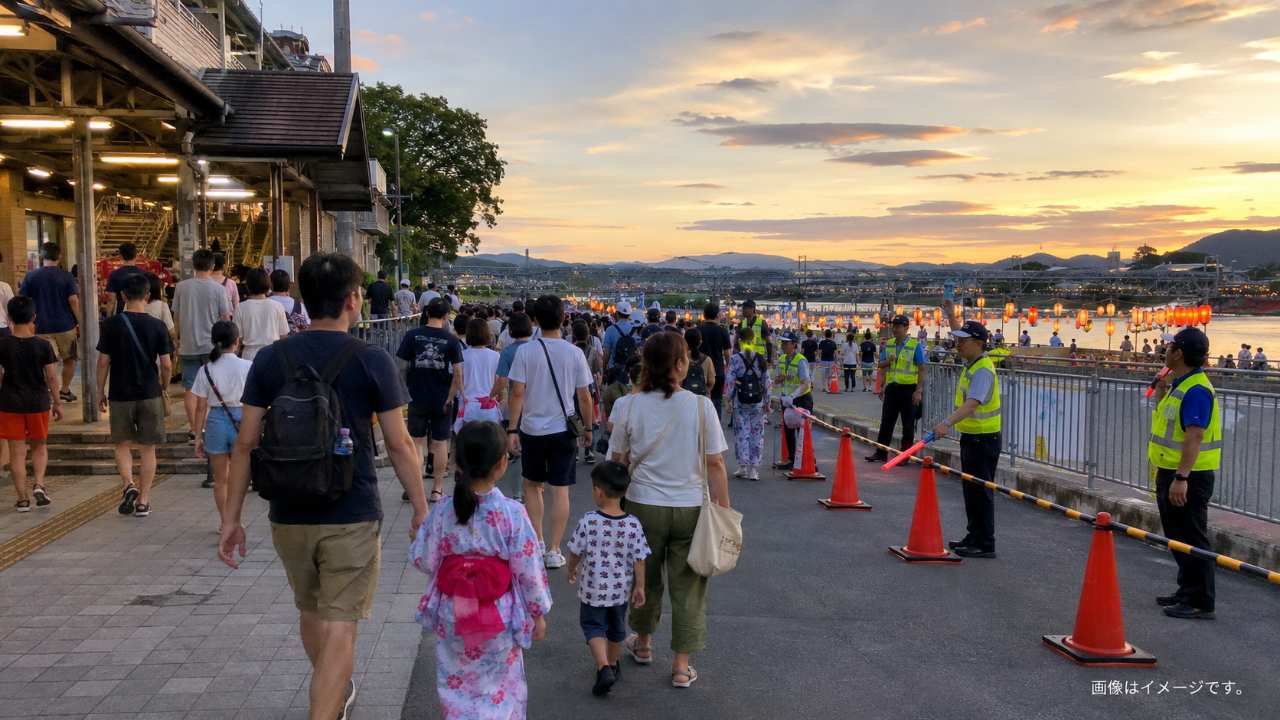 夏祭り当日の地方駅周辺をイメージした実写写真風。夕方の駅前から河原の会場へ向かう家族や友人たちが、係員の案内に従って歩いている