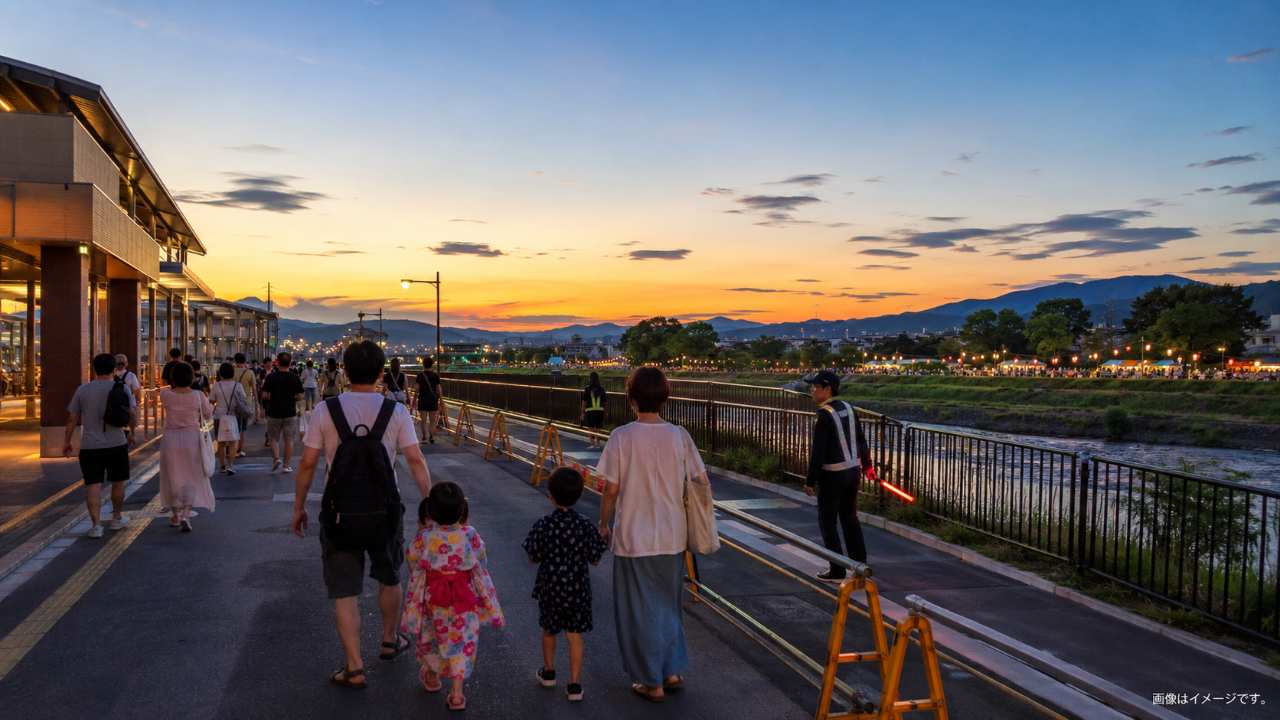 夏祭りの夕方、郊外の鉄道駅東口から川沿いの祭り会場へ歩く家族や友人たちを描いた実写写真風